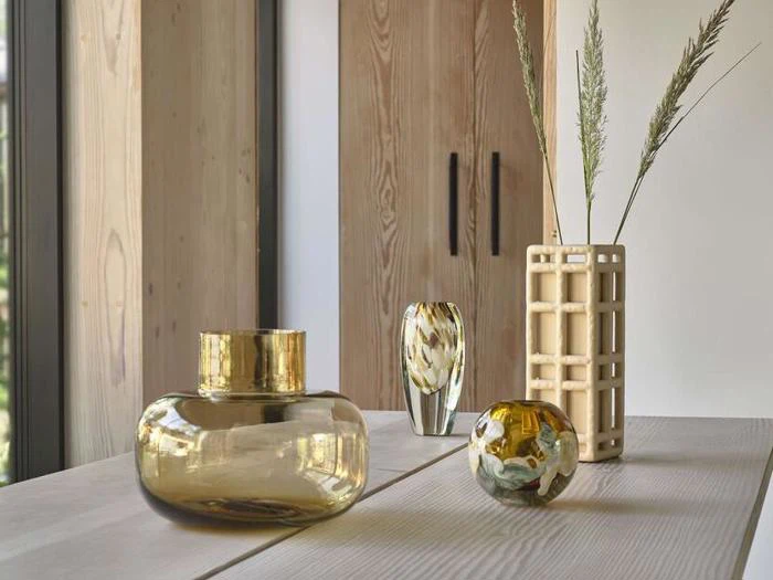 På et moderne bord står en guldkrukke, en lille glaskugle, en vase med tørret græs og et højt glasornament i et minimalistisk rum med træskabe.