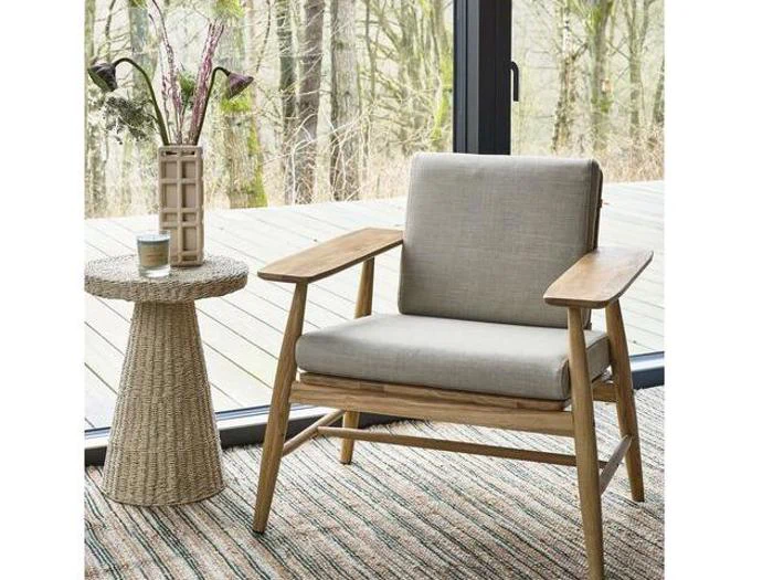 En trælænestol med grå hynder står ved siden af et flettet sidebord med en vase med blomster og en kop i nærheden af store vinduer med udsigt over en skov.