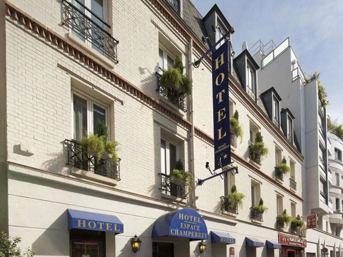 Gadebillede af et lyst hotel med blå skilte med teksten HOTEL ESPACE CHAMPERRET, med små balkoner med potteplanter og flere vinduer.