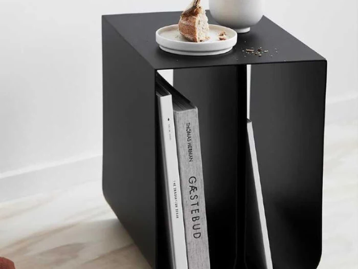 Et moderne sort sidebord har en tallerken med brød og en kop på toppen; bøger opbevares lodret i en indbygget rille nedenunder.