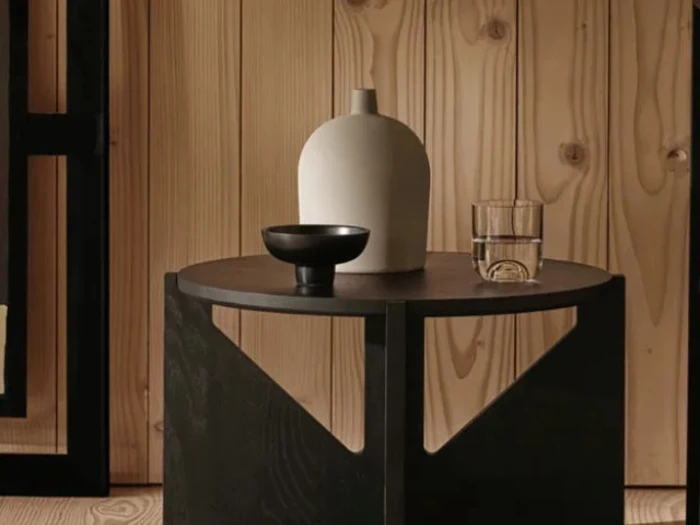 På et rundt sidebord af træ står en hvid keramikvase, en sort skål og et glas vand op ad en væg med træpaneler.