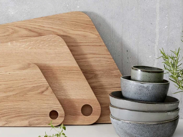 To skærebrætter af træ og en stak med fire grå keramikskåle står på et køkkenbord op ad en betonvæg. Nogle grønne krydderurter står i nærheden.