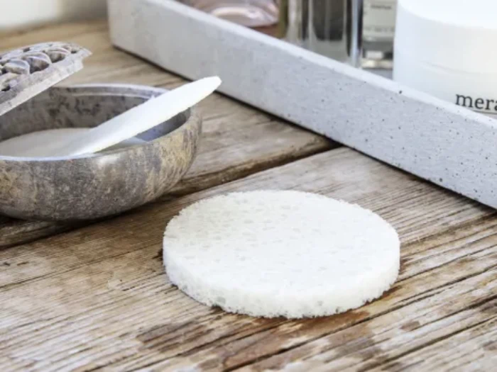 En rund, hvid rensesvamp ligger på en træplade ved siden af en grå skål med en spatel og en hvid bakke med hudplejeprodukter.