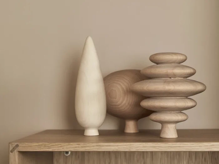 Tre abstrakte træskulpturer med glatte, minimalistiske former står på en lys træhylde mod en beige baggrund.