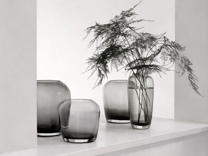 Fire gennemskinnelige grå glasvaser er arrangeret på en hvid hylde, den ene med sarte grønne blade, mod en minimal hvid baggrund.