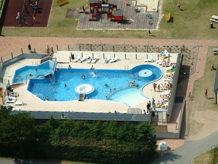 Luftfoto af en swimmingpool med folk, der svømmer og slapper af, omgivet af et solbadningsområde; en legeplads og græs er synlige i nærheden.