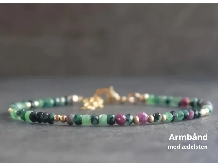 Et delikat ædelstensarmbånd med små grønne, rosa og guldfarvede perler på en grå overflade. Teksten lyder: Armbånd med ædelsten.