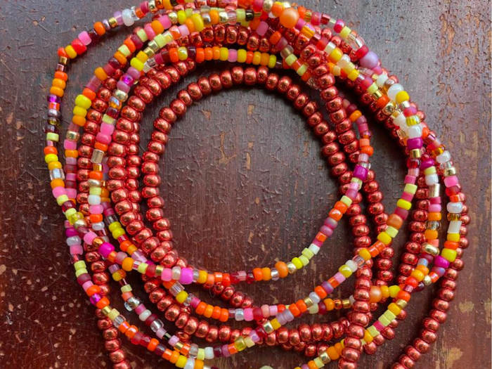 Flere farverige perlekæder med røde, orange, gule, pink og grønne perler er viklet sammen på en mørk træoverflade.