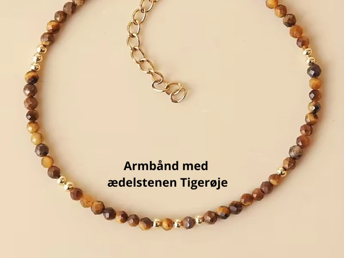 Et perlearmbånd med brune og gyldne tigerøje-ædelsten og en guldkædeforlænger, vist på en beige baggrund. Dansk tekst: Armbånd med ædelstenen Tigerøje.