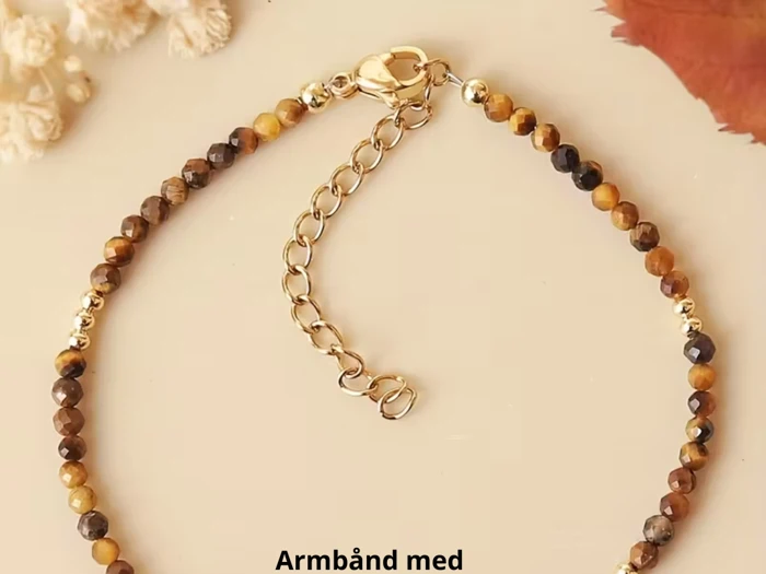 Et perlearmbånd med guld og brune sten, en guldkædeforlænger og en lås, vist på en lys overflade.