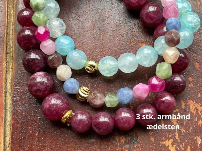 Tre perlearmbånd med ædelsten i forskellige farver, herunder rødbrun, pink, grøn, blå og guldaccenter, udstillet på en træoverflade. Teksten lyder 3 stk. armbånd ædelsten.