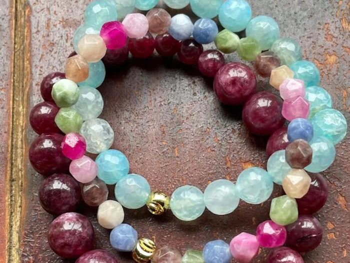 Nærbillede af flerfarvede perlearmbånd med runde og facetterede perler i røde, blå, grønne, rosa og gyldne nuancer på en rustik brun overflade.