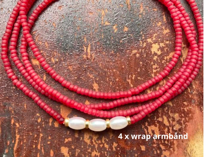 Nærbillede af et wrap-armbånd i fire lag med små røde perler og tre hvide ovale perler, placeret på en rustik brun overflade. Teksten lyder 4 x wrap-armbånd.