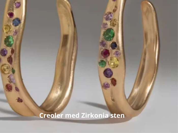 To guldøreringe dekoreret med farverige zirkonia-sten på en neutral baggrund. Teksten i bunden lyder: Creoler med Zirkonia sten.