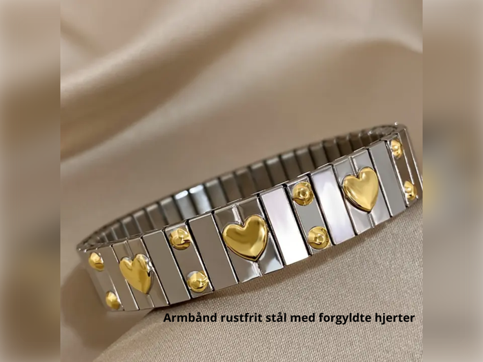 Et sølvfarvet armbånd i rustfrit stål med guldbelagte hjerter og prikker på en beige stofbaggrund. Tekst: Armbånd rustfrit stål med forgyldte hjerter.
