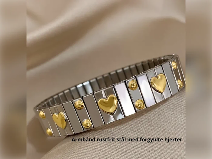 Et sølvfarvet armbånd i rustfrit stål med guldbelagte hjerter og prikker på en beige stofbaggrund. Tekst: Armbånd rustfrit stål med forgyldte hjerter.