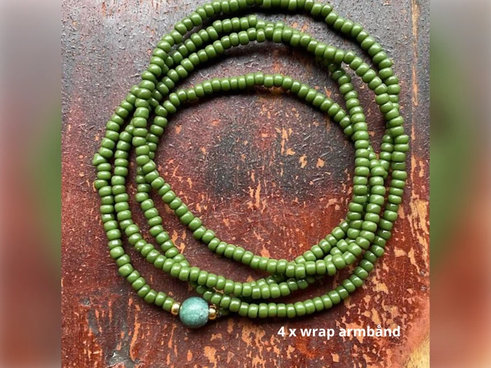 Et grønt perlearmbånd med guldaccenter og en enkelt blå perle, viklet i fire omgange, på en træoverflade. Teksten lyder 4 x wrap-armbånd.