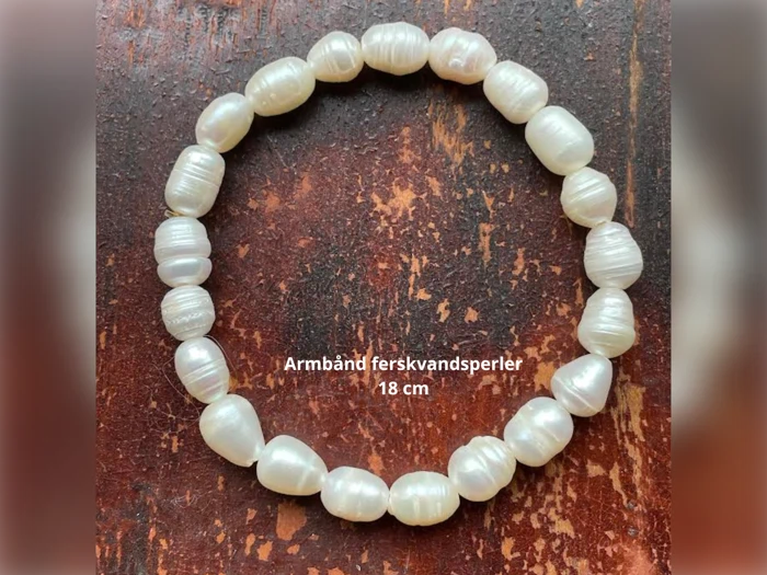 Et armbånd af hvide ferskvandsperler arrangeret i en cirkel på en træplade, mærket Armbånd ferskvandsperler 18 cm.