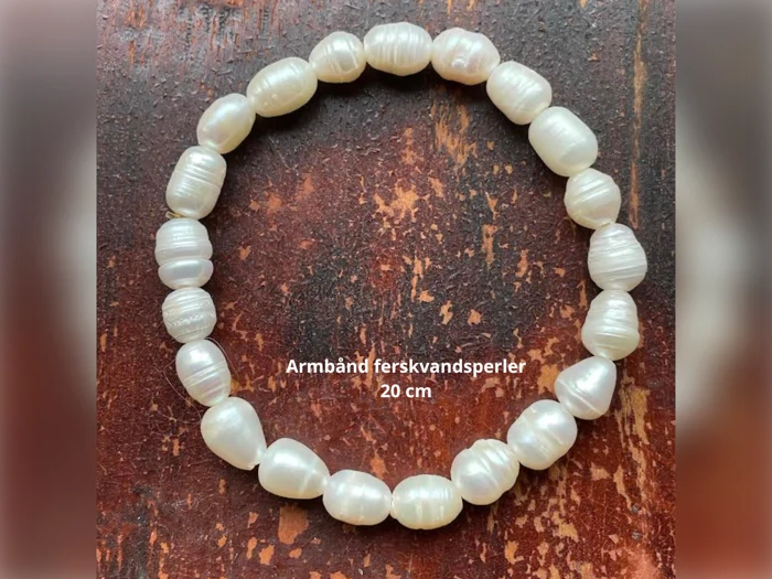 Et armbånd lavet af hvide ferskvandsperler arrangeret i en cirkel på en træoverflade, med teksten Armbånd ferskvandsperler 20 cm i midten.