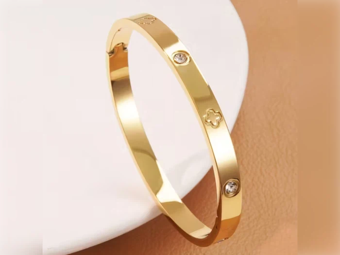 Et guldarmbånd med små diamantlignende sten og kløverformede indgraveringer på en hvid og beige baggrund.