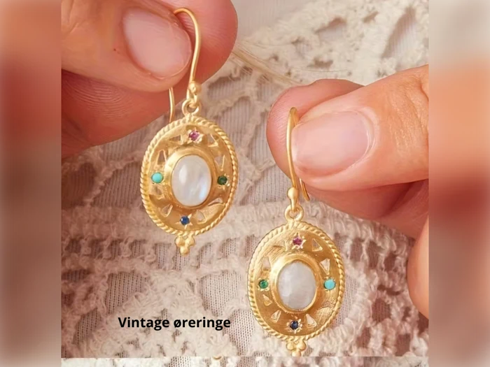 To hænder holder udsmykkede guldøreringe med ovale perlecentre, farverige ædelstensaccenter og indviklede detaljer. Teksten Vintage øreringe er synlig under øreringene.