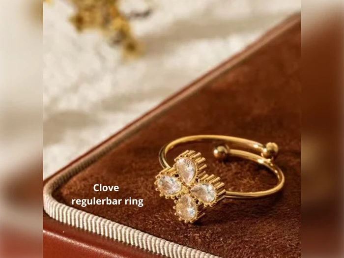 En guldring med et firkløver-design, prydet med klare ædelsten, placeret på en brun struktureret overflade. Teksten lyder: Nellike regulerbar ring.