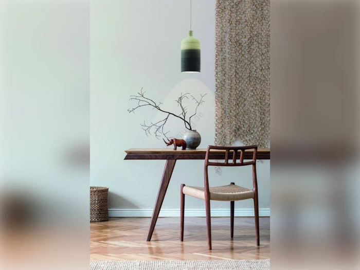 En minimalistisk spiseplads med et træbord, en stol, en vase med nøgne grene, en lille pyntegenstand og en vævet pendel over. Neutrale toner og hyggelig indretning.