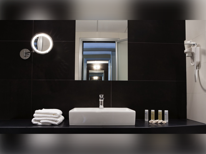 Et moderne badeværelse med en firkantet håndvask, stablede håndklæder, toiletartikler, en væghængt hårtørrer og et rundt, oplyst spejl foran et stort spejl, der reflekterer rummet.