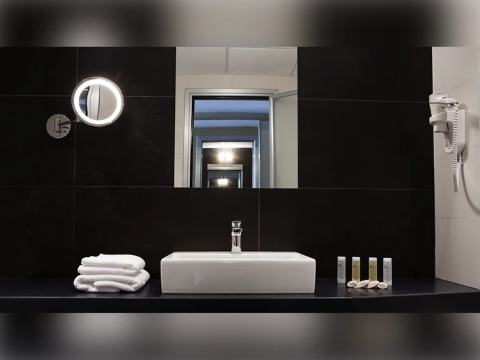 Et moderne badeværelse med en firkantet håndvask, stablede håndklæder, toiletartikler, en væghængt hårtørrer og et rundt, oplyst spejl foran et stort spejl, der reflekterer rummet.