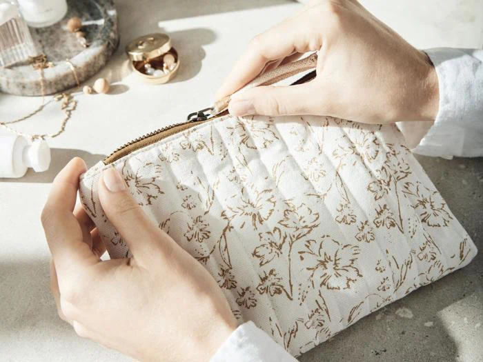 En persons hænder holder en cremefarvet stofpose med et guldfarvet blomstermønster og lyner den op på en lys overflade med smykker og kosmetik i nærheden.