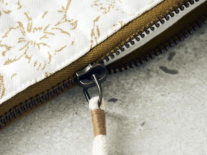 Nærbillede af en metallynlås på et stofstykke med blomstermønster og en snoretrækflig, der ligger på en lysegrå struktureret overflade.