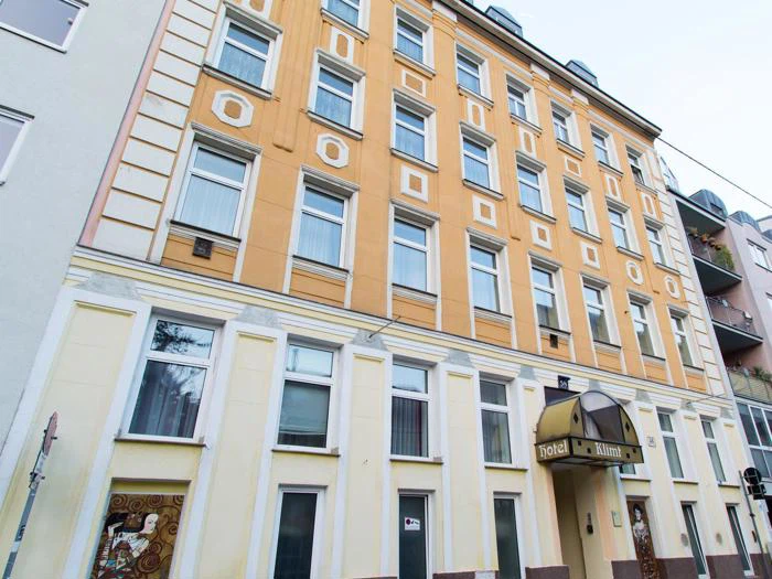 En bygning i flere etager med gule og beige vægge, store vinduer og et skilt med teksten "hotel" over indgangen. Facaden har dekorative arkitektoniske detaljer.