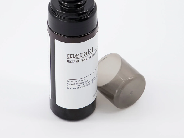 En sort flaske mærket meraki Instant Tanning Mousse med en grå hætte placeret ved siden af på en hvid baggrund.