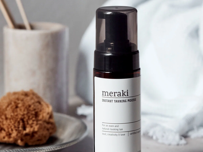 En flaske Meraki Instant Tanning Mousse med sort hætte står på en lys flade ved siden af en naturlig svamp og et hvidt håndklæde i baggrunden.