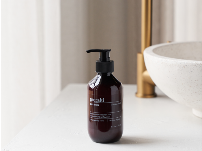En mørk pumpeflaske med Meraki bodylotion står på en hvid bordplade i nærheden af en rund, hvid vask og en messingvandhane.