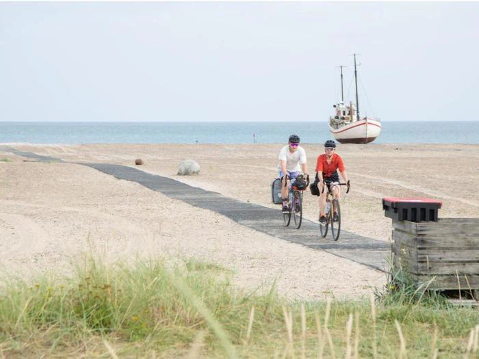 To cyklister kører langs en sandet strandsti med en båd på bredden og havet i baggrunden under en lys himmel.