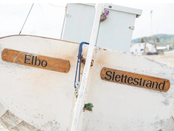Stævnen af en hvid træbåd på en strand, med navnene Elbo og Slettestrand skrevet på træplader.