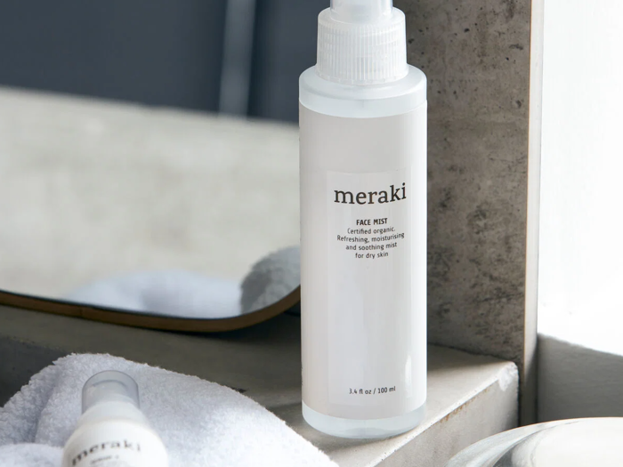 En hvid flaske med påskriften meraki FACE MIST står på et badeværelsesbord nær et spejl, og et hvidt håndklæde og en anden flaske er delvist synlige i nærheden.