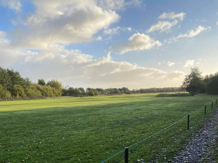En bred græsmark omkranset af træer under en delvis overskyet himmel med sollys, der bryder igennem, og et rebhegn, der løber langs en grussti.