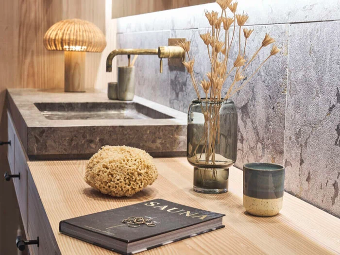 En moderne badeværelsesbordplade med stenvask, messingarmatur, tørrede blomster i en glasvase, en svamp, en keramisk kop, en lampe og en bog med titlen SAUNA.