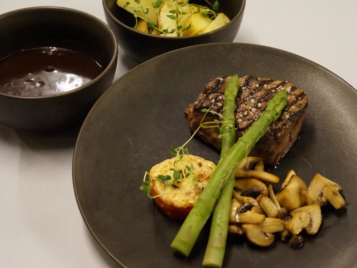 Et tallerkenmåltid med grillet bøf toppet med asparges, ristet tomat, sauterede svampe, en skål brun sauce og en salat med agurk og mikrogrønt ved siden af.