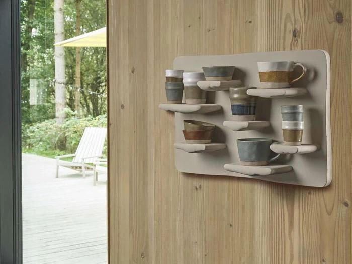 Ni forskellige keramiske kopper står på en moderne væghængt hylde, der er fastgjort til en trævæg med et vindue, der viser en udendørs terrasse og træer.