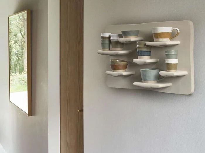 Et væghængt display med forskellige keramiske kopper og krus på buede hylder, placeret i et moderne, minimalistisk interiør.