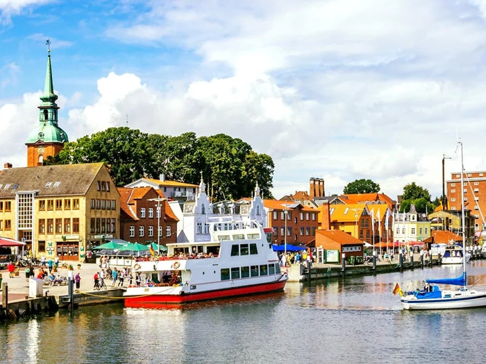 Farverig havnefront med både, der ligger til kaj, folk, der går langs promenaden, og charmerende europæiske bygninger, herunder en kirke med et grønt spir, under en delvist overskyet himmel.
