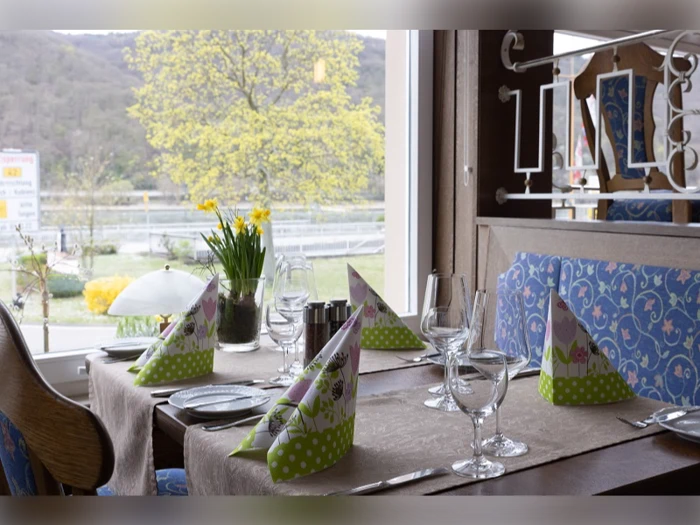 Et restaurantbord dækket til fire med foldede servietter, vinglas og en vase med gule blomster ved et vindue med udsigt til træer og en flod.