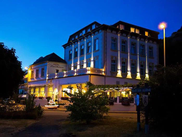 Et veloplyst hotel med klassisk arkitektur står under en dybblå aftenhimmel, omgivet af træer og buske, og en gadelampe lyser i nærheden.