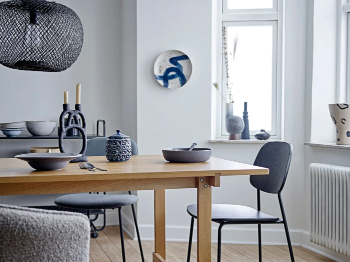 En moderne spisestue med træbord, sorte stole, keramisk service og minimalistisk indretning i neutrale toner. Lyset strømmer ind gennem de høje vinduer og lyser rummet op.