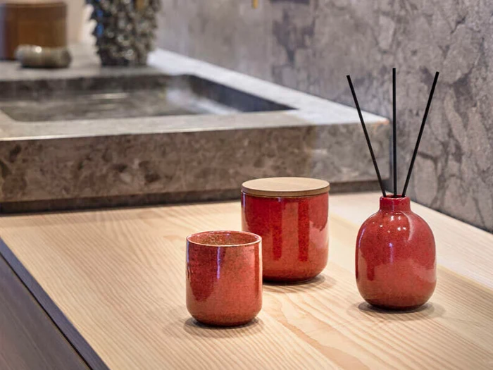 Røde keramikbeholdere og en sivdiffusor er placeret på en lys træoverflade i nærheden af en grå stenvask i et moderne badeværelse.