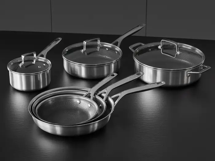 Et sæt køkkengrej i rustfrit stål, bl.a. tre stegepander og tre gryder med låg, anbragt på en sort bordplade.