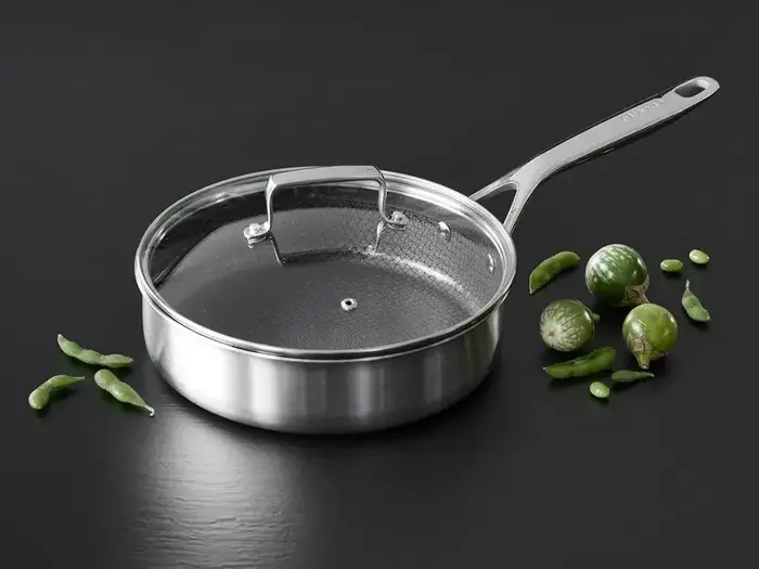 En sauterpande i rustfrit stål med glaslåg står på en mørk flade ved siden af grønne ærtebælge og små grønne auberginer.